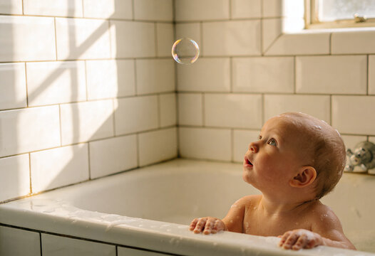 Curious baby sitting in bathtub gazing up at floating soap bubble, childhood wonder and bath time play with warm natural light