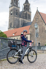 Obraz premium Young girl helmet stands bicycle cobblestone bridge historic european Magdeburg city cathedral towers background. Urban lifestyle scene childhood independence safe cycling travel atmosphere