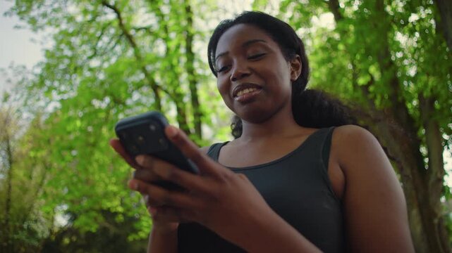 African American woman looking at smartphone and smiling while reading message creating positive and uplifting outdoor lifestyle moment in park environment