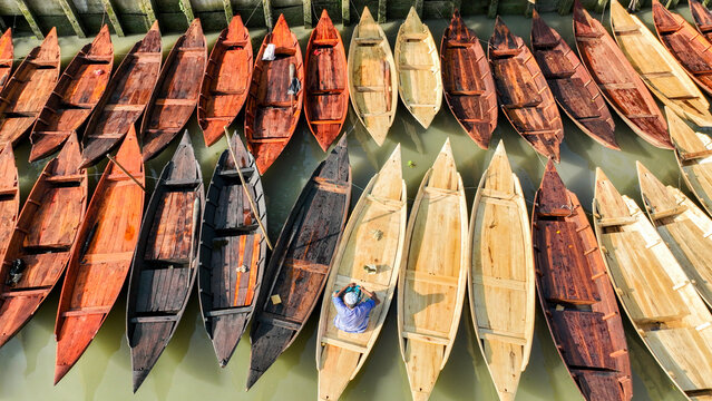 Aerial view of a fleet of wooden boats, arrayed in a spectrum of earthy hues, resting on the still, olive-green waters of Pirojpur Pourashava, Pirojpur Pourashava, Barisal Division, Bangladesh.