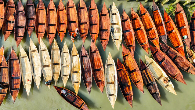 Aerial view of an array of wooden boats with rich, warm tones contrasting with the cool, green water, Pirojpur Pourashava, Barisal Division, Bangladesh.