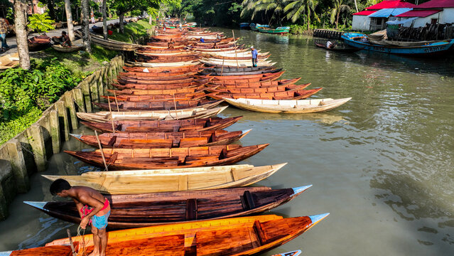 Aerial view of a long line of wooden boats, their prows gleaming under the sun, moored along the canal's edge, Pirojpur Pourashava, Barisal Division, Bangladesh.