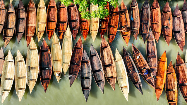 Aerial view of boats aligned on the river, their wooden hulls displaying a spectrum of browns and creams against the river's green surface, Pirojpur Pourashava, Barisal Division, Bangladesh.