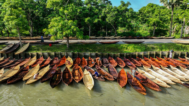 Aerial view of wooden boats aligned on the riverbank contrasting with the lush green trees along the road, Pirojpur Pourashava, Barisal Division, Bangladesh.