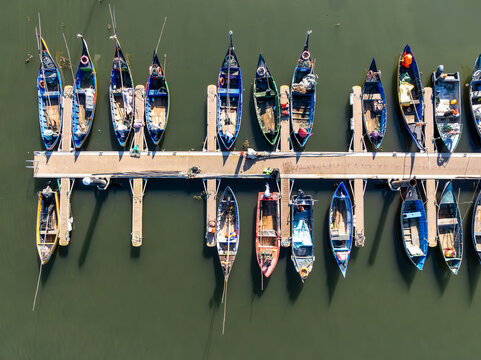 Aerial view of fishing boats, berthed in a row, casting elongated shadows on the dark water in Porta de Abrigo da Torreira, Torreira, Aveiro, Portugal.