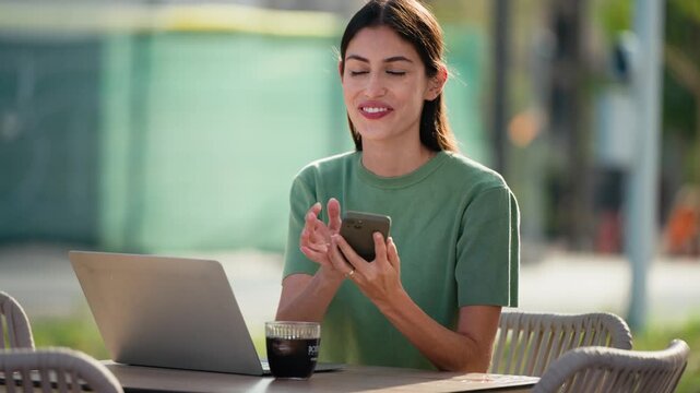 Video of happy businesswoman working with laptop while talking with smartphone having a drink on a terrace in the city