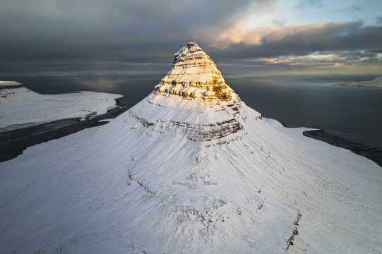 Aerial view of snow-covered Kirkjufell mountain basks in the soft glow of the setting sun, contrasting sharply with the dark, moody sky above, Kirkjufell, Grundarfjardarbaer, Iceland.