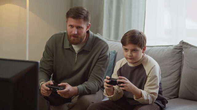 Medium long shot of focused adult Caucasian father sitting on sofa next to gen Alpha son playing video games with controllers in front of TV in living room