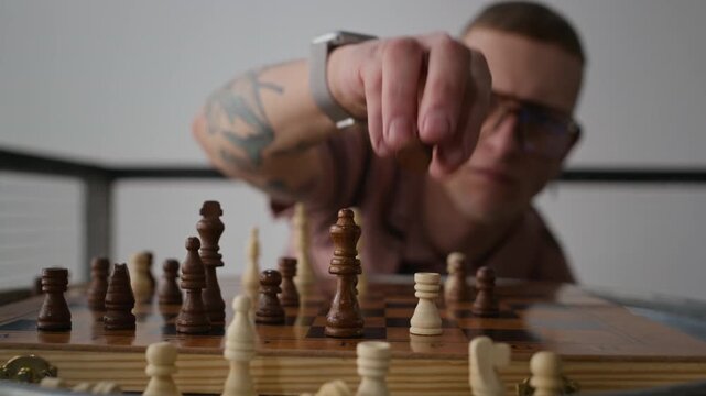 Closeup shot of focused tattooed Caucasian man in glasses thinking before making move while playing chess game indoors, developing skill and logic