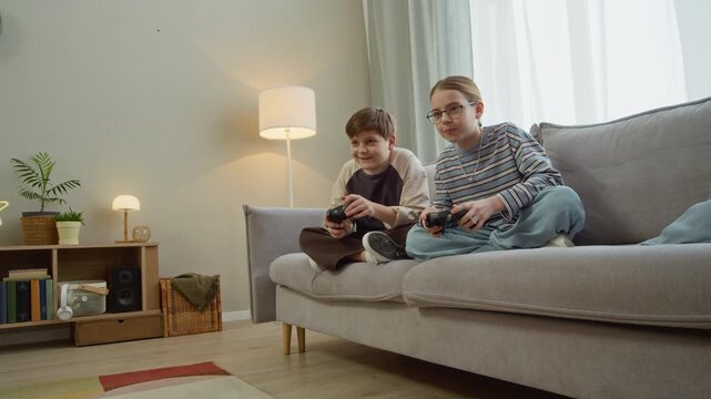 Wide shot of two Caucasian gen Alpha kids playing video games with joysticks while sitting on sofa in cozy living room, copy space