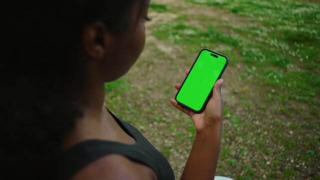 African American woman holding smartphone with green screen while seated outdoors creating mockup and template ready mobile device usage moment in natural environment