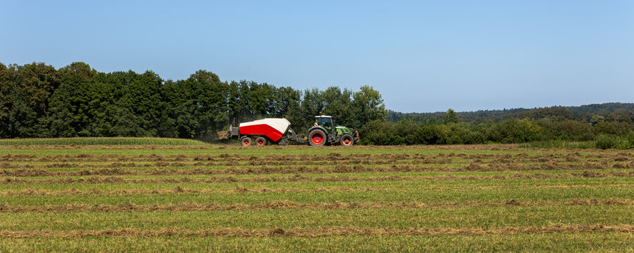 Side view of big modern tractor machine with baler trailer making hay blocks after wheat field harvesting. Agricultural equipment and agribusiness machinery