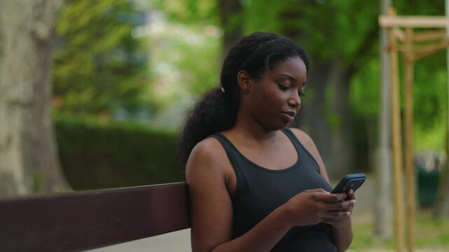 African American woman seated on park bench using smartphone and looking down at screen with calm and focused expression creating quiet and natural outdoor lifestyle moment in green environment