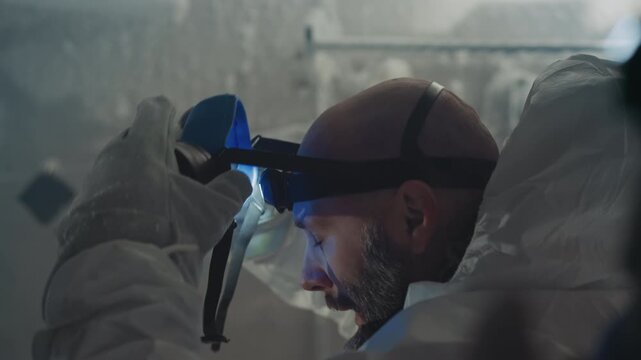 White researcher in full respirator looking upward hooded hazmat suit frost on beard headlamp beam illuminates icy pipes quiet resolve preparing instruments sample collection under nuclear winter