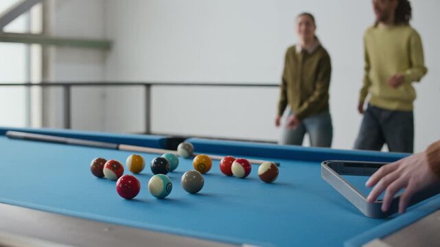 Medium shot of multiethnic group of friends setting up billiard balls in triangle formation on blue pool table, getting ready to play