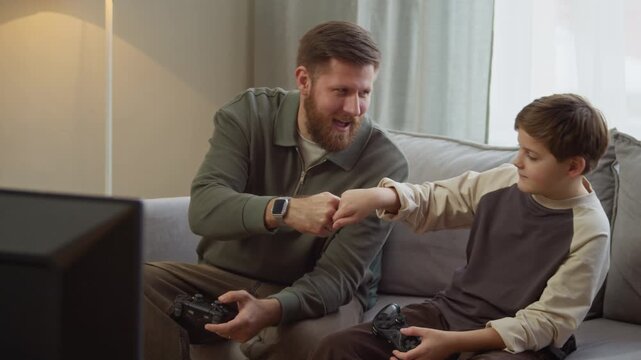 Medium long shot of adult Caucasian father fist bumping with young son sitting on sofa while playing video games with joysticks at home