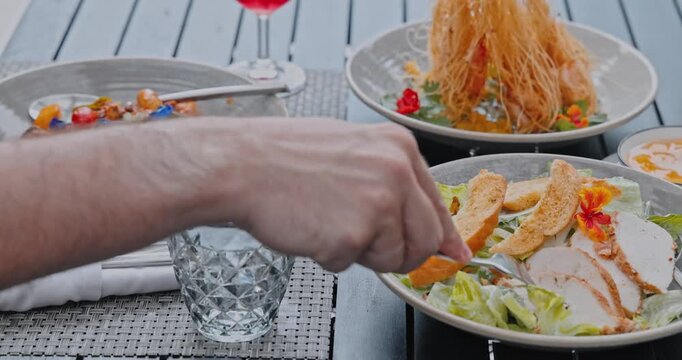 Hand picking piece of sliced chicken with fork, Caesar salad and Asian fusion dishes served on outdoor restaurant table, summer lifestyle, restaurant menu advertising