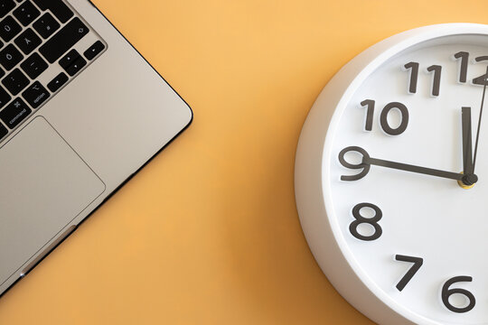 Close‑up of a laptop and wall clock on a pastel orange background, symbolizing productivity, time management, remote work and modern workspace organization.