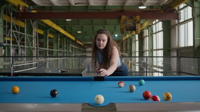 Medium eye level stab shot of young Caucasian woman focusing and lining up shot with cue on billiards table in industrial loft environment, showing concentration and precision