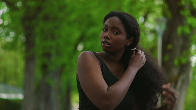 African American woman standing outdoors adjusting her hair with both hands creating natural personal care moment in green park environment