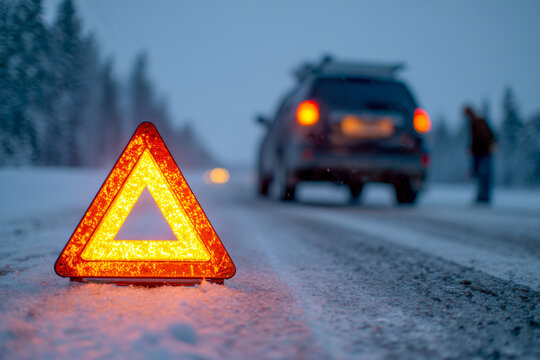 Reflective emergency warning triangle placed on a snowy road with a stopped vehicle and a person standing beside it during dusk in winter forest surroundings