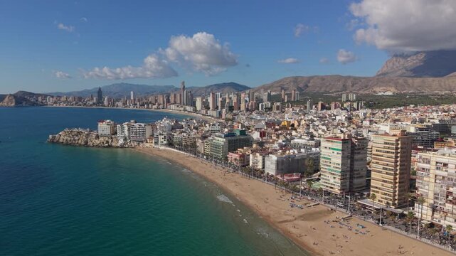 Benidorm Costa Blanca Spain high drone flight towards Benidorm Castle headland with views of both Levante and Ponientes beaches with calm sea,mountain background fluffy clouds blue sky and bright sun