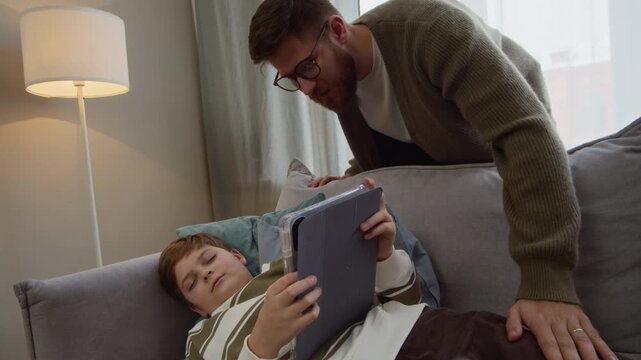 Handheld shot of Caucasian dad taking digital tablet from hands of gen Alpha son sleeping on sofa and waking up then