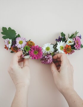 Top view of hands delicately arranging a colorful floral garland on a beige background, perfect for DIY wedding d&eacute;cor, spring celebration invitations, and lifestyle floristry blogs