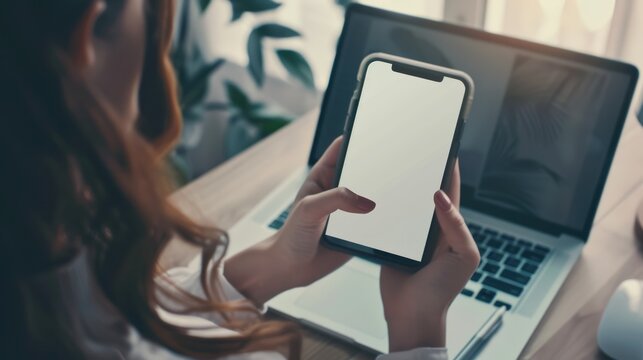 Woman Holding Smartphone in Front of Laptop in Bright Workspace
