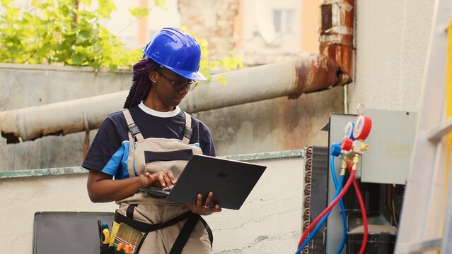 Expert repairman servicing external HVAC system internal components, checking freon level and calibrating thermostats. Proficient worker imputing maintenance results data on laptop