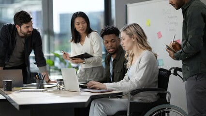 Obraz premium Diverse group of young professionals collaborates around a laptop in a bright modern office, including a blonde woman in a wheelchair, while reviewing a whiteboard marketing plan.