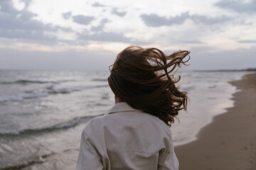 Obraz premium A cinematic rear view of a young woman standing on beach, her hair blowing wildly in the strong wind. The overcast sky and moody ocean waves create a feeling of peaceful solitude, raw nature, freedom
