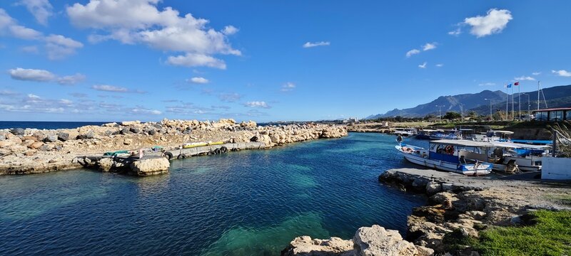 Panoramic view of Lapta fishing harbor with turquoise waters and stone piers. A peaceful Mediterranean scene featuring boats, mountain backdrops, and a cloudy blue sky along the rocky coastline.