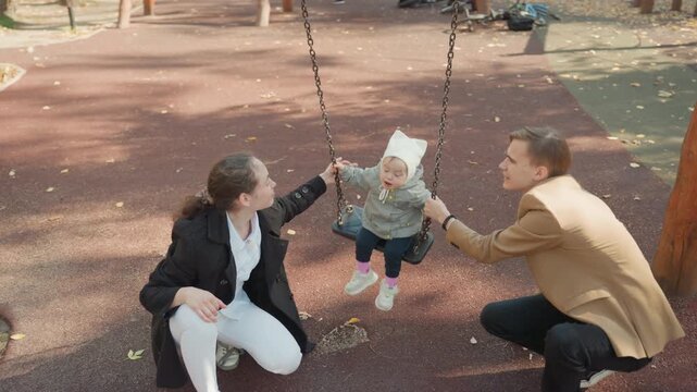 Parents pushing toddler on swing at playground, young married couple gently play with baby in sunny autumn park, protective hands steady toddler, warm smiles, casual jackets and hat, stroller