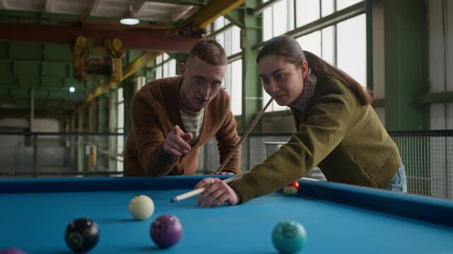 Eye level medium shot of young Caucasian woman focusing and aiming cue ball while playing billiards with male friend at blue pool table in indoor setting
