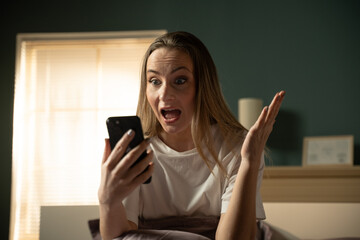 Fototapeta premium Woman in bedroom reacting with disbelief and astonishment while reading shocking news or an unexpected message on her smartphone, conveying emotional impact and unexpected information