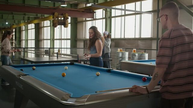 Medium wide shot of two couples enjoying indoor games in spacious industrial loft, one couple playing billiards in foreground and another playing table tennis in background