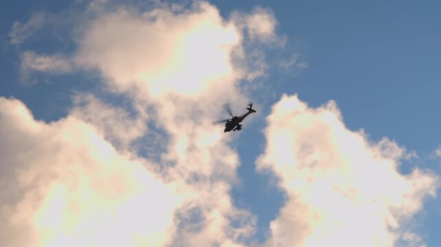 Military attack helicopter flying in slow motion through a cloudy blue sky. High angle silhouette of a combat aircraft patrolling with rotating blades during a daytime mission.