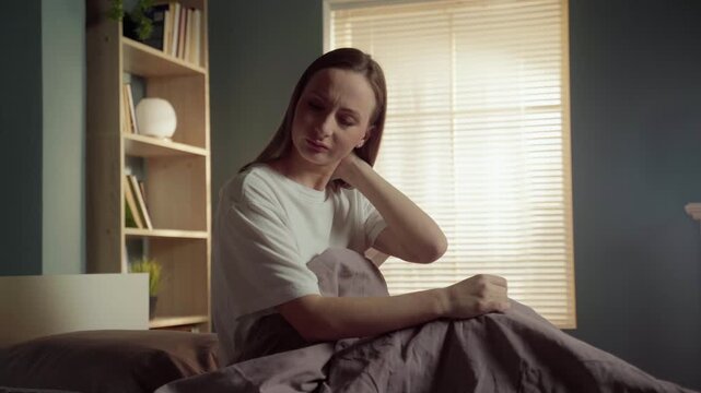 Uncomfortable young woman sitting in bed after waking up, suffering from neck pain and muscle stiffness, massaging the sore spot and grimacing from discomfort in her modern sunlit bedroom