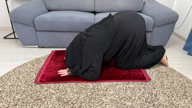 Muslim woman in black abaya performing sujud prostration on a red prayer mat at home