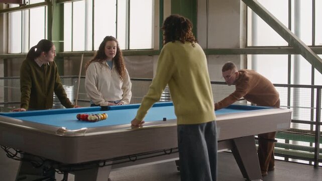 Medium wide shot of four diverse friends gathering around blue pool table and man making break shot, beginning game together