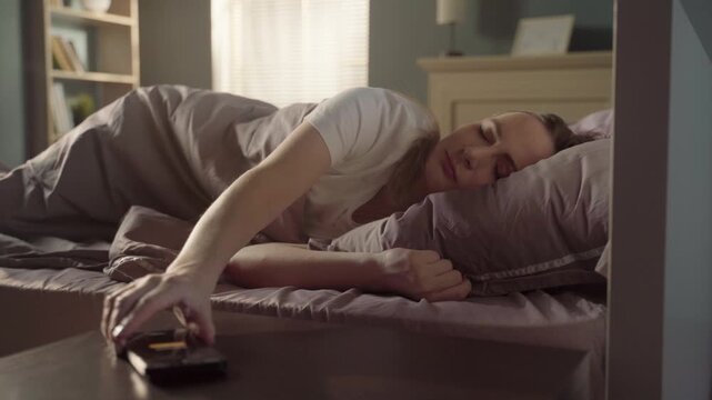 Young woman sleeping in her bed being awakened by the sound of her smartphone alarm, she reaches out to turn it off and continues to rest, annoyed by the early wake up call