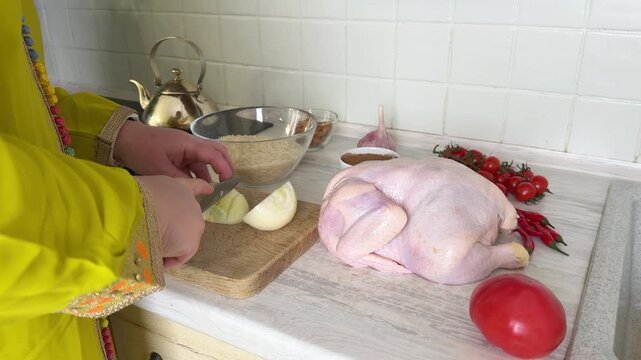 Close up of a person in a yellow dress slicing a white onion on a wooden board. A whole raw chicken, rice, tomatoes, and spices are on the kitchen counter, ready for cooking a meal