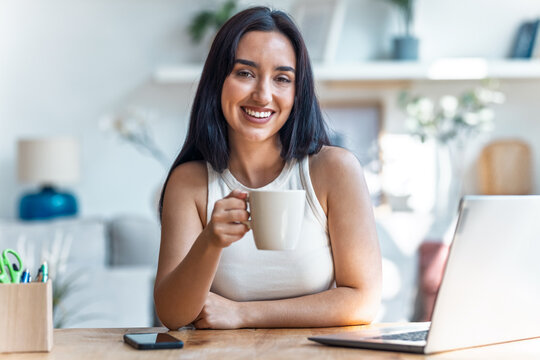 Pretty woman looking at camera while drinking a cup of coffee working with laptop in the livingroom at home