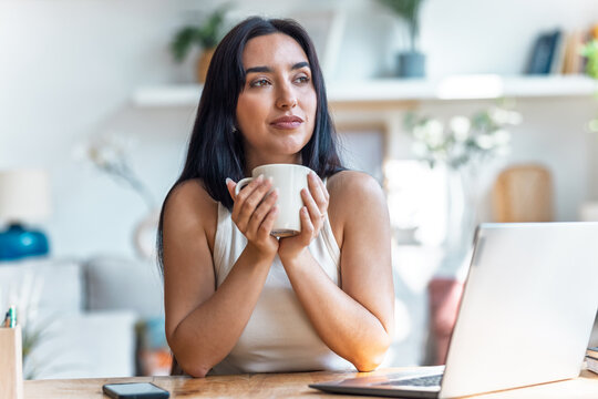 Pretty woman drinking a cup of coffee while working with laptop in the livingroom at home