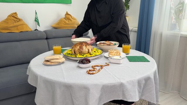 A woman in a black abaya sets table for a festive dinner. She serves a traditional Middle Eastern meal with roasted chicken, rice, dates, and bread. A Saudi Arabian flag is in background