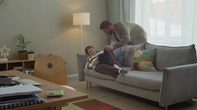 Wide shot of Caucasian father waking up gen Alpha son sleeping with digital tablet in hands on sofa in cozy living room