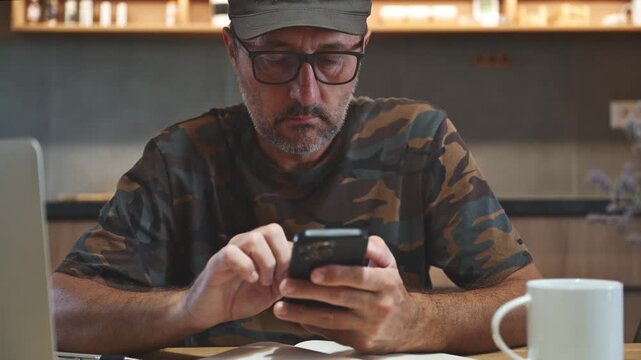 Male wearing army hat and military camouflage shirt drinking coffee and using mobile phone in the morning in modern indoor setting, 4K