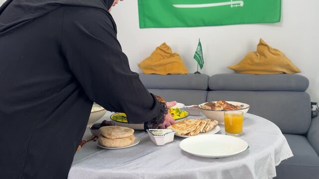Person in black abaya places dish of roasted chicken and rice onto table set for traditional Middle Eastern feast. Saudi Arabian flags decorate room, suggesting celebration or holiday