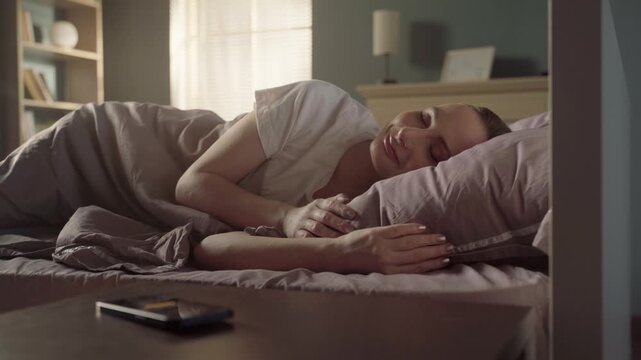 Peaceful young woman waking up in her cozy bed in the sunlit bedroom, smiling and stretching after a restful sleep before turning off her smartphone's alarm in the morning